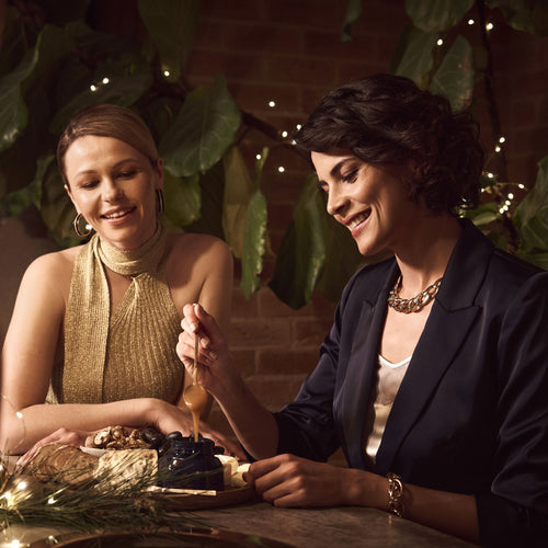 Two women sitting at a table with a decorative setting, smiling and interacting with the Manuka Health Manuka Honey MGO 600+ Kit.