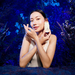 Woman holding a skincare product against a blue, nature-themed background.