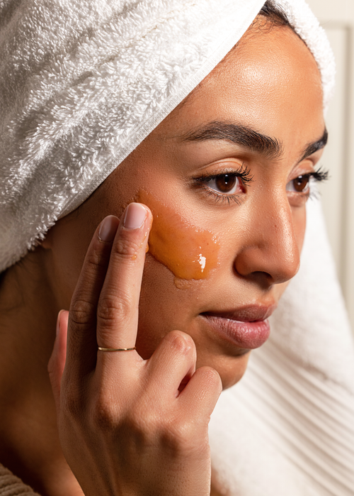 Woman applying a manuka honey facial mask to her face with a towel on her head.