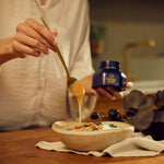 Manuka Health Manuka Honey with person drizzling honey from a spoon into a bowl with a jar of honey in the background.