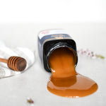 Manuka Health Manuka Honey jar with honey spilling out on a white surface with a wooden honey dipper.