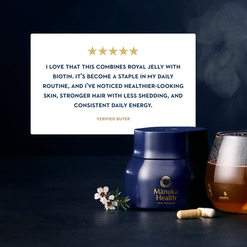 A dark blue jar of Manuka Health New Zealand cream sits on a dark surface with a glass of amber liquid, a small flower sprig, and a capsule. A large white review card with gold stars and text is positioned behind the jar.